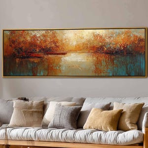 Puede incluir: Una pintura de paisaje rectangular con marco dorado. La obra representa una serena escena lacustre con árboles en tonos naranja y rojo. El agua refleja los colores de los árboles y el cielo, creando una atmósfera tranquila. La pintura se exhibe sobre un sofá.