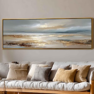 Puede incluir: Pintura abstracta de paisaje en un marco dorado. La obra representa una escena costera serena con una paleta de colores apagados de beige, gris y dorado. La pintura se exhibe sobre un sofá de madera con varios cojines.