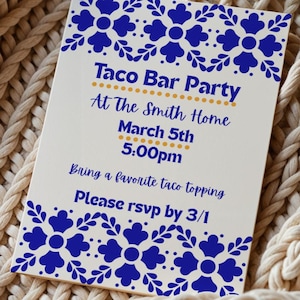 Puede incluir: Una tarjeta de invitación rectangular con fondo blanco y diseños florales azules. El texto "Taco Bar Party" se muestra en dorado, con detalles del evento en azul. La tarjeta está sobre una manta de punto beige.