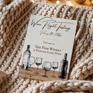 May include: A cream-colored card with the text "Wine Flight Tasting" and event details. The card features illustrations of wine bottles and glasses. The card is resting on a beige, chunky knit blanket.