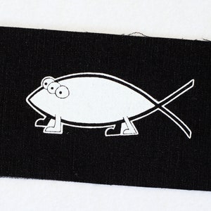 May include: A black fabric patch with a white outline of a fish with three eyes and legs. The fish is walking on its legs.