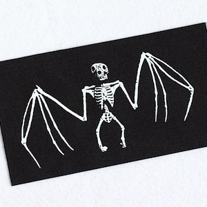May include: A rectangular black fabric patch featuring a white screen-printed graphic of a bat skeleton. The design displays the skull, rib cage, and extended wing bones of the creature centred on the dark background with clean, sharp lines.