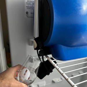 Puede incluir: Un aparato azul y negro está montado dentro de un refrigerador. Se sostiene una copa de vino transparente con texto blanco frente al aparato. El texto en el vaso dice "FRESH FRESH FRESH".