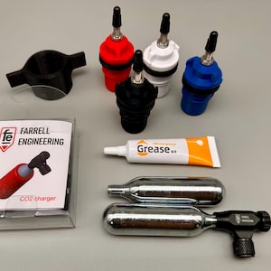 May include: A CO2 charger kit including a clear plastic box labeled Farrell Engineering, a black plastic handle, four colored valve adapters in red, white, blue, and black, a tube of silicone grease, and two silver metal CO2 cartridges.