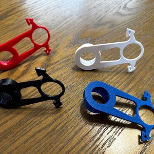 May include: Four colorful, 3D-printed bottle openers in red, white, black, and blue. Each opener features a circular grip, a rectangular opening, and a pointed end. The openers are arranged on a wooden surface.