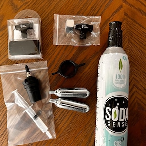 May include: An assortment of Soda Sense products, including a CO2 cylinder, black plastic components, and small silver cartridges. The Soda Sense cylinder is white with a black label and the words "Soda Sense" and "CO2".