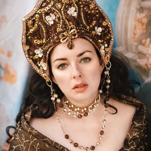 Peut inclure: Une femme porte une coiffe marron ornée de détails dorés et blancs, de perles et de motifs floraux. Elle porte également un collier assorti et un châle marron avec un motif de feuilles dorées. L'ensemble est somptueux.