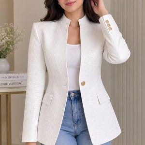 Op de afbeelding: Een witte, getextureerde blazer met een opstaande kraag, lange mouwen met drie gouden knopen bij de manchetten en een enkele gouden knoopsluiting in de taille. Het jasje heeft twee klepzakken en wordt gedragen over een wit topje en een blauwe spijkerbroek.