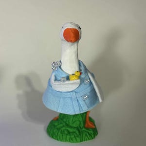 Puede incluir: Una figura caprichosa de ganso con cabeza y cuello blancos, pico naranja y patas naranjas. Viste un abrigo azul claro con ribete blanco y sostiene un pequeño patito amarillo. La figura se apoya sobre una base de hierba verde.