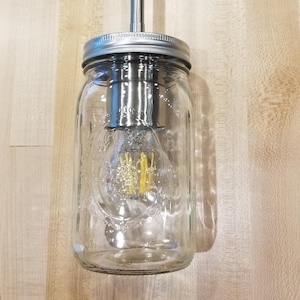 May include: A clear glass mason jar pendant light with a silver metal lid and a silver rod. The light bulb inside is visible and lit. The light fixture hangs against a light wood background.