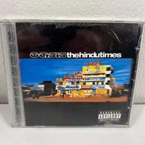 Könnte beinhalten: Eine CD-Hülle mit dem Album "the hindu times" von Oasis. Das Cover zeigt ein farbenfrohes Gebäude vor blauem Himmel. Der Bandname ist weiß, mit einem Hinweis auf die Eltern.