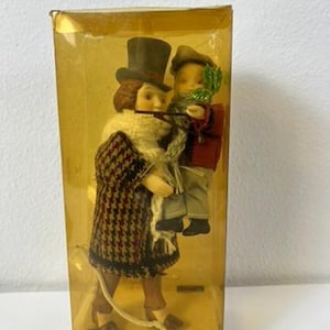 Puede incluir: Figurita decorativa dentro de una caja de plástico transparente. La figurita representa dos figuras, una con sombrero de copa y abrigo estampado, la otra con vegetación. La caja tiene una base dorada con el texto "Santas World".