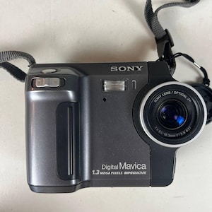 May include: A dark gray Sony Digital Mavica camera with a lens and attached strap. The camera has a flash and the text "SONY" is visible. The lens has the text "SONY LENS / OPTICAL 3x".