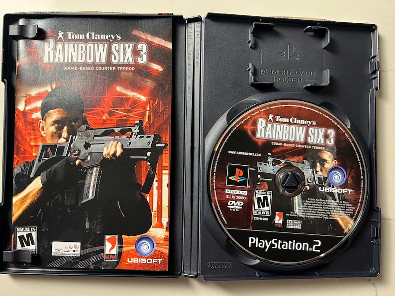 Puede incluir: Caja de juego de PlayStation 2 abierta para Tom Clancy's Rainbow Six 3. La car&aacute;tula presenta a una persona sosteniendo un arma. El disco del juego es visible dentro de la caja.