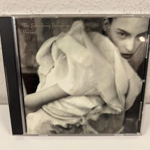 May include: A CD case featuring the album "Perfect" by The Smashing Pumpkins. The cover art is a black and white image of a person holding a white cloth. The text "The Smashing Pumpkins" and "Perfect" are visible.
