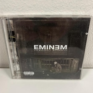 Puede incluir: Una funda de CD con el álbum "The Marshall Mathers LP" de Eminem. La portada es oscura con el nombre del artista en letras blancas. Se muestra una advertencia para padres.
