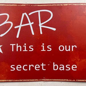 May include: Red metal sign with white text that reads "BAR", a star, and "This is our secret base". The sign has a distressed look with a slightly worn appearance, suggesting a vintage or rustic style.
