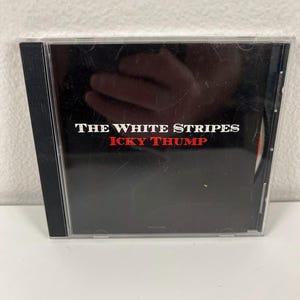May include: A compact disc case with a black cover. The text "THE WHITE STRIPES" is printed in white, and "ICKY THUMP" is in red. The case is clear plastic with a black spine.