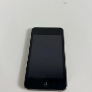 May include: A black digital media player with a rectangular screen and a single square home button centered at the bottom of the front panel. The device features a slim, rounded metallic frame surrounding the dark display area.