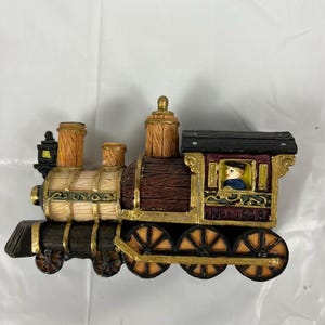 May include: A decorative train figurine with a detailed design. The train features a brown and gold color scheme, with accents of black and cream. The wheels are gold, and the train has a small figure in the cabin. The text "NO. 1938" is visible.