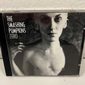 May include: A black and white CD case featuring the album "Zero" by The Smashing Pumpkins. The cover art is a grayscale portrait of a person with short, light-colored hair. The band's name and album title are printed in white.