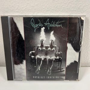 May include: A CD case with the album cover for Jane's Addiction's "Nothing's Shocking." The artwork shows two nude figures with flames above their heads, seated in a rocking chair. The band name and album title are displayed.