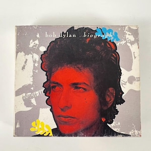 May include: Album cover for Bob Dylan's "Biograph." The cover features a red-toned portrait of Dylan with black hair, set against a grey background with guitar silhouettes. The album title is visible.