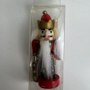 May include: A wooden nutcracker figurine in a clear plastic case. The nutcracker has a red hat, gold crown, and a long white beard. It is wearing a red and black uniform with gold accents. The base is red.