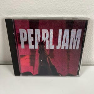 Puede incluir: Una caja de CD con una portada roja y negra con el nombre de la banda "PEARL JAM" en letras plateadas grandes. La portada incluye una silueta oscura de una persona. La caja del CD está en una funda de plástico transparente.