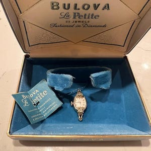 May include: An open vintage Bulova La Petite watch box with a blue velvet interior. The box contains a small, ornate wristwatch with a white face and a blue velvet band. A small tag reads "BULOVA La Petite". The box is trimmed in gold.