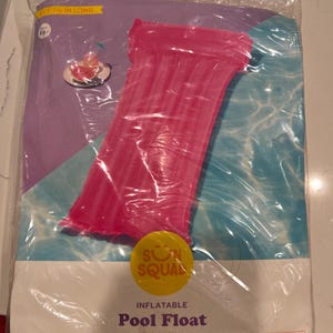 Op de afbeelding: Een roze opblaasbaar zwembad drijver in een doorzichtige plastic verpakking. De verpakking heeft de woorden "Pool Float" en "Built-in pillow" erop gedrukt. Er is ook een gele cirkel met de woorden "Sun Squad".