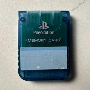 May include: A blue PlayStation memory card with the PlayStation logo and the words "PlayStation" and "MEMORY CARD" printed on it. The card has a light blue label on the front and is made of translucent plastic.