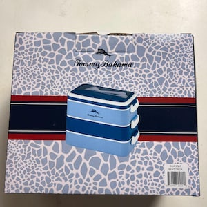 May include: A Tommy Bahama bento box, stacked in three sections, is displayed on a patterned box. The bento box is blue and white with a logo on the top. The box has a light blue giraffe-like pattern with red and navy blue stripes.