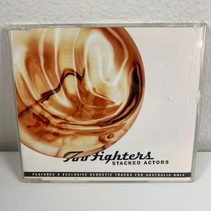 May include: A CD case featuring the Foo Fighters' album "Stacked Actors." The cover art displays a swirling, abstract design in shades of brown and tan. The band's name and album title are printed in black, with additional text at the bottom.