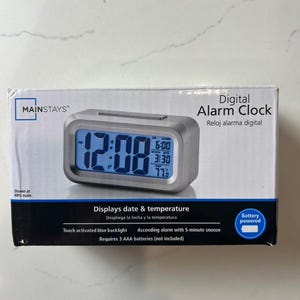May include: A digital alarm clock by Mainstays, with a silver frame and a blue digital display showing the time, date, and temperature. The clock features a touch-activated blue backlight and requires 3 AAA batteries. The box reads "Digital Alarm Clock".