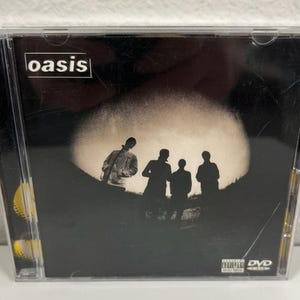 Könnte beinhalten: Eine CD-Hülle der Band Oasis. Der Name der Band steht in Weiß auf schwarzem Hintergrund. Die Abbildung zeigt Silhouetten vor einer großen, hellen, kreisförmigen Lichtquelle.