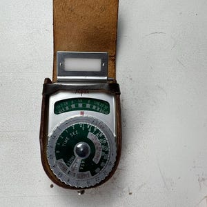 May include: Vintage Aipex light meter in a brown leather case. The meter has a silver face with black and green markings, including f-stop, time, and ASA settings. The leather case has a flap and a belt loop.