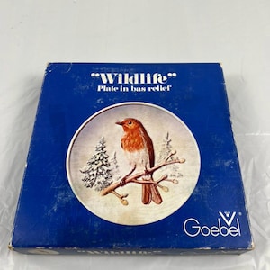 May include: A blue box with a decorative plate featuring a red-breasted bird perched on a branch. The plate is in bas-relief and has a winter scene with evergreen trees. The box reads "Wildlife" and "Goebel".