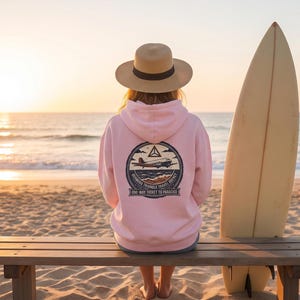 May include: A light pink hooded sweatshirt featuring a circular graphic on the back with an airplane illustration, the text Bermuda Triangle Travel Agency, and the phrase One-Way Ticket to Paradise, paired with a tan wide-brimmed sun hat.