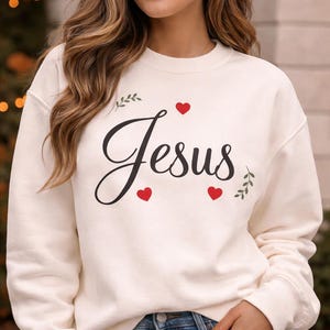 Peut inclure: Sweat-shirt crème avec le mot "Jesus" en écriture cursive, accompagné de cœurs rouges et de brins. Le haut à manches longues est de style décontracté, adapté au quotidien.