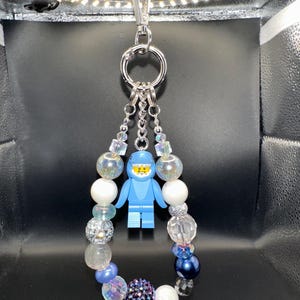 May include: A handmade keychain featuring a blue Lego figure in a spacesuit design. The keychain includes a silver clasp, a large silver ring, and a beaded charm with white, iridescent, and blue beads. The charm is approximately 4 inches long.