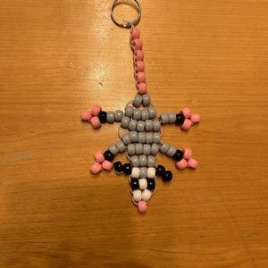 May include: A beaded opossum keychain. The opossum is made of gray, black, white, and pink beads. The keychain has a silver ring and a pink beaded chain. The opossum has its arms and legs spread out.