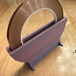 May include: A roll of copper tape sits in a purple and blue tape dispenser. The copper tape is a bright, metallic color, and the dispenser has a simple, functional design. The dispenser is made of a solid material and has a rectangular shape.