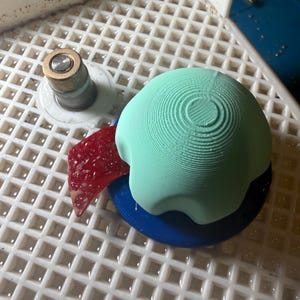 Op de afbeelding: Een lichtgroen, bolvormig object met concentrische cirkelvormige ribbels staat op een blauwe basis. Een rood, driehoekig stuk en een metalen cilinder zijn ook zichtbaar. De objecten staan op een wit, rasterpatroon oppervlak.