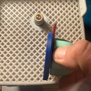 Op de afbeelding: Een close-up van een glassnijgereedschap met een blauwe handgreep en een rood mes. Het gereedschap bevindt zich op een wit plastic rooster. Het gereedschap wordt door een hand vastgehouden.