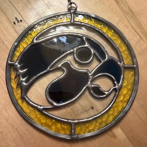 May include: Stained glass suncatcher featuring the Iowa Hawkeyes logo, a black and gold hawk head within a circular frame. The glass has a textured, iridescent finish, and is framed with a metal border and hanging chain.