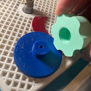 Op de afbeelding: Een verzameling glas snijgereedschap en accessoires. Inclusief een blauw cirkelvormig onderdeel, een rood gebogen stuk, een lichtgroene knop en een kleine metalen cilinder. De items zijn gerangschikt op een wit rooster oppervlak.