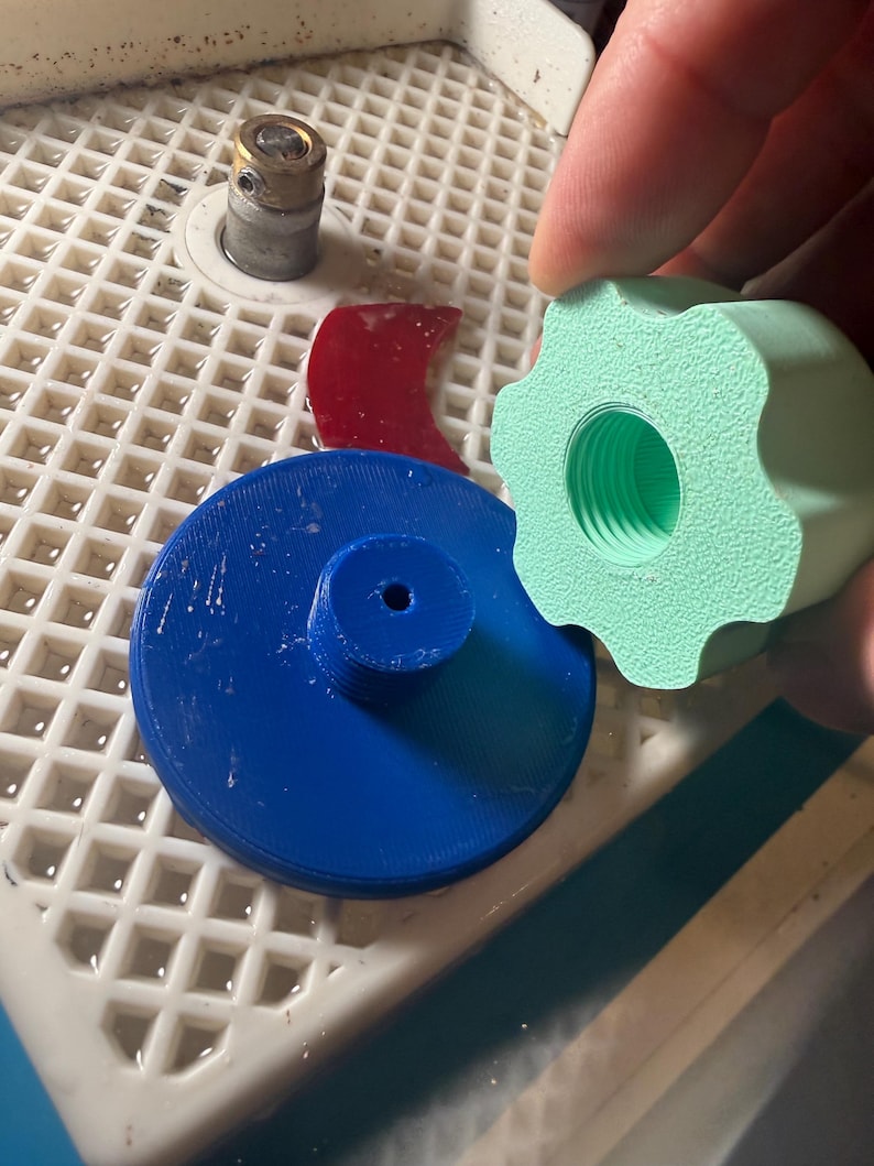Op de afbeelding: Een verzameling glas snijgereedschap en accessoires. Inclusief een blauw cirkelvormig onderdeel, een rood gebogen stuk, een lichtgroene knop en een kleine metalen cilinder. De items zijn gerangschikt op een wit rooster oppervlak.