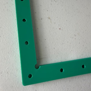 May include: A green, L-shaped plastic frame component featuring a series of evenly spaced circular holes along its length. The corner of the frame includes a small, circular cutout, and the surface exhibits a subtle, textured finish.