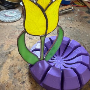 May include: A stained glass tulip with a bright yellow flower head and two light green leaves, featuring dark metal solder lines. The glass piece is inserted into a circular, purple, segmented base with a radial pattern of grooves.